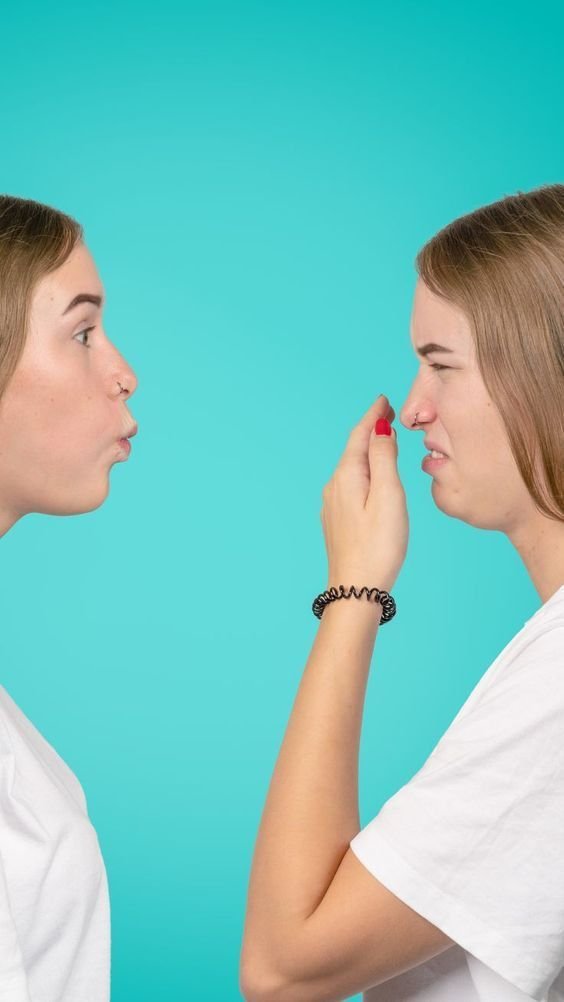 Two people facing each other, one blowing air and the other covering their nose with an uncomfortable expression.